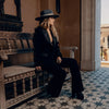 Woman in a black outfit and hat sitting on a wooden bench in an ornate setting.