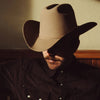 Man wearing a cowboy hat and dark shirt with a mustache, sitting indoors.