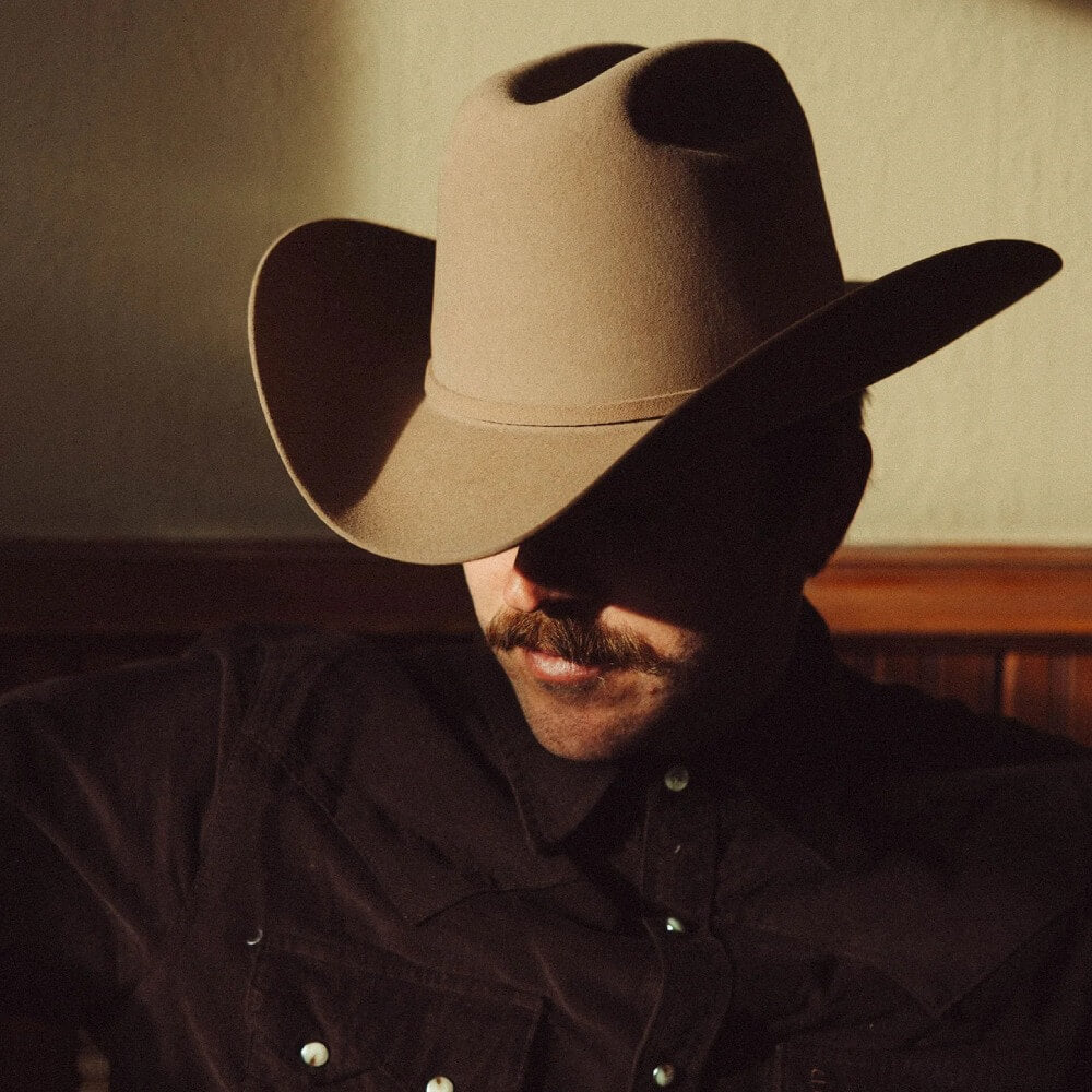 Man wearing a cowboy hat and dark shirt with a mustache, sitting indoors.