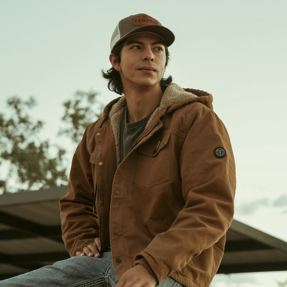 Man wearing a brown jacket and cap outdoors with trees in the background