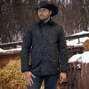 Man wearing a black jacket and cowboy hat standing in a snowy outdoor setting with hay bales.