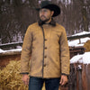 Man wearing a tan coat and black cowboy hat standing in a snowy outdoor setting with hay and wooden structures.