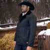 Man wearing a black coat and cowboy hat standing in a snowy rural setting with hay bales and wooden fences.