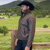 Man wearing a brown jacket and black cowboy hat standing in a green field with mountains in the background