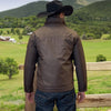 Man wearing a brown leather jacket and cowboy hat standing in a scenic landscape with mountains and green fields.