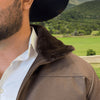 Man wearing a brown jacket with fur collar and white shirt, standing outdoors with green landscape in the background