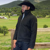 Man wearing a black jacket and cowboy hat standing in a scenic outdoor setting with green fields and mountains.