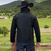 Man wearing a black jacket and cowboy hat standing in a scenic landscape with mountains and green fields.
