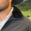 Person wearing a dark jacket with a fur collar, standing outdoors with greenery in the background