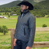 Man wearing a gray jacket and black cowboy hat standing in a green field with mountains in the background