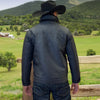 Man wearing a black jacket and cowboy hat standing in a scenic landscape with mountains and green fields.
