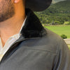 Person wearing a dark jacket with a fur-lined collar, standing outdoors with greenery in the background.