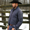 Man wearing a navy puffer jacket and black cowboy hat standing in front of a wooden fence with snow on the ground.