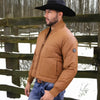 Man wearing a brown puffer jacket and black cowboy hat standing in front of a wooden fence with snow on the ground.