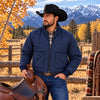 A man wearing a navy blue quilted zip-up jacket, blue jeans, and a black cowboy hat stands outdoors in a rural landscape with mountains, autumn trees, and a wooden fence, holding a brown leather saddle.