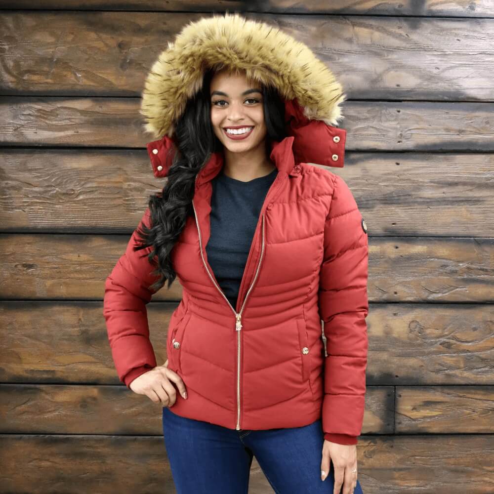 Woman wearing a red puffer jacket with a fur-lined hood against a wooden wall.