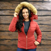 Woman wearing a red puffer jacket with a fur-lined hood against a wooden wall.